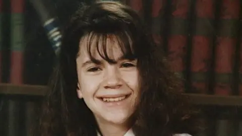 A school photo of caroline glache. She is a little girl of about 14 with long dark brown hair. She is smiling directly on camera. There is a bookshelf behind him.