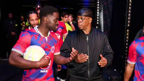 Getty Images Ian Wright walks through the tunnel alongside a player dressed in a blue and red kit and holding a football in one arm. The two look as if they are locked in a deep discussion.
