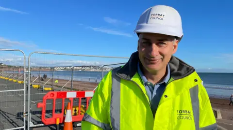 A man with a Torbay Council hard hat and jacket stands in front of the area where they are building the new flood defences on Paignton seafront.