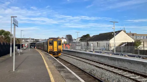 Network Rail A picture of Newquay station from the platform. A GWR train is moving away. There is a white building on the platform opposite. There is a Newquay sign on the platform the picture is taken from and a bench. The sky is blue with white clouds. 