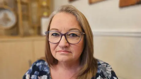 Darlin's shoulder length has golden hair and is wearing a floral navy top. She is looking at the camera directly and wearing glasses.