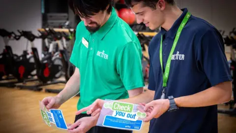 GLL Two leisure centre workers wearing blue and green polo tops hold stickers reading 'Check You Out'