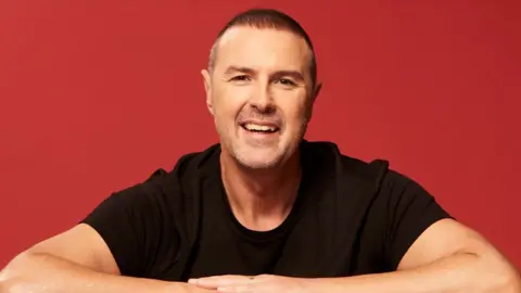 Paddy McGuinness smiling at the camera while leaning on a table with his arms folded in front of him, against an orange/red background. He has short dark hair and is wearing a black t-shirt.