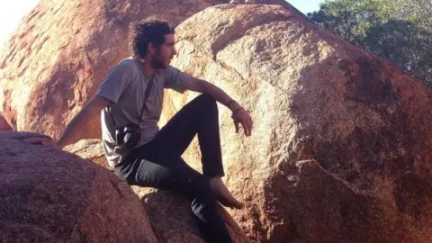 FAMILY HANDOUT Adam Cornell sitting on a large boulder, with two other boulders behind him. He is looking to the right and wearing a grey t-shirt and black trousers, with a camera over his shoulder. He has dark hair and a beard. 