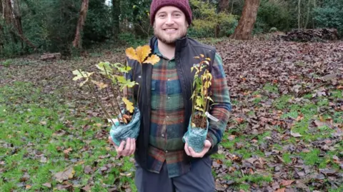 Leicestershire County Council A man wearing a purple hat and colourful shirt holding two tree saplings. He is standing in an area that has lots of trees behind him