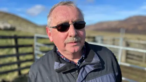 A man wearing dark glasses, a checked shirt and dark jacket in a sheep pen on a hillside.