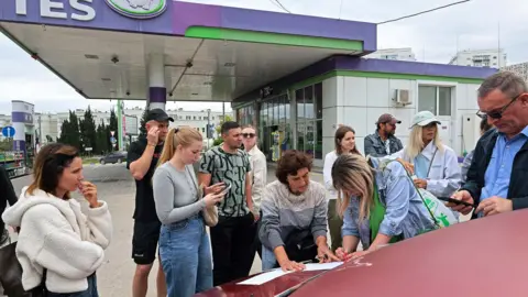 People dressed in casual clothing at a petrol station in Sevastopol, Crimea leaning on a car bonnet putting their names on a list to join the queue for fuel after the authorities restricted fuel sales amid a supply shortage following Ukrainian attacks on logistics routes in the course of Russia-Ukraine conflict, June 1, 2026. 