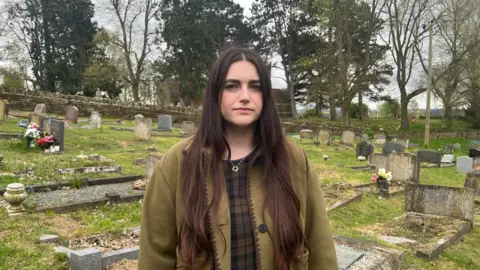 Tony Fisher/BBC A woman with long black hair and wearing a brown coat standing in a cemetery with gravestones in the background. She is wearing a necklace and has a sad expression on her face.