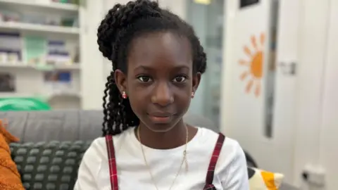 Kiishi is looking directly at the camera with a blurred background. She has black skin, brown eyes and long hair tied up. She is wearing red earrings and a gold necklace