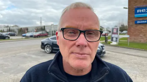 John Devine/BBC Glen Hindmarsh, 61, has short strawberry blonde hair and has dark rimmed glasses on he is wearing a dark blue fleece top with a dark shirt beneath. Behind him is a road with a black car passing others cars are parked across the road and a business is on the left