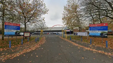 Google A road leads up to a sports complex in the distance. Signs on either side of the road read "Tipton Sports Academy".