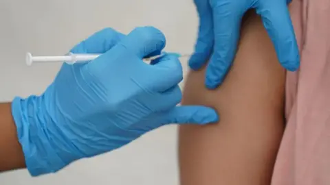 PA Media A person wearing blue gloves injects a syringe into the arm of another person wearing a pink top.