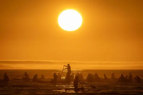 Andre Coelho/EPA-EFE People sitting or standing on paddle boards in the ocean with the sun shining overhead in a bright yellow sky