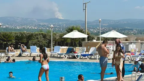 People sunbathe and swim in a hotel pool with smoke visible in the distance 