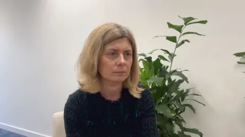 Deputy Lyndsay Feltham is wearing a blue dress and has blonde hair. She is sat in front of a plant. 