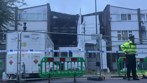 A police officer stands in front of a fence, that has been erecting a the front of a terraced property gutted by fire.