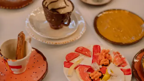 Teacups and saucers and a plate showing a breakfast that are all made of clay. The breakfast plate seems to have clay bacon, mushrooms and beans on it.