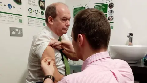 Sir Michael McBride sitting down as he receives his flu jab. He has short dark hair and is wearing a white shirt with a green tie. The jab is being administered by a pharmacist wearing a pink striped shirt and short dark hair.