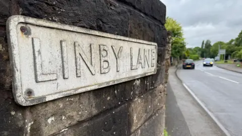 Street sign of Linby Lane, in Linby, Nottinghamshire, with two cars on the road to the right.