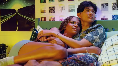 Netflix/Heartstopper Season 3 A scene from Hearstopper with Elle and Tao hugging on Tao's bed. Elle is resting her head on Tao's chest and they're staring at something off-camera. Both of them are wearing pyjamas and are lying on top of the covers.