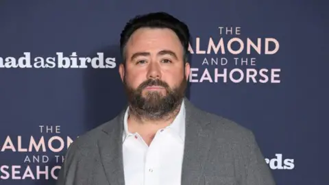 Getty Images Celyn Jones wearing a grey blazer and a white shirt. He is standing in front of a dark blue screen. He has short dark hair and a short beard. 