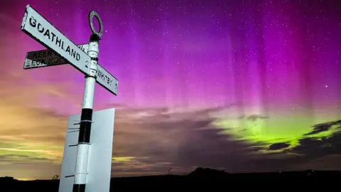 The Northern Lights light up the sky behind a black and white signpost