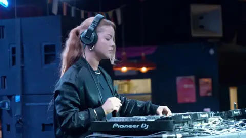 Harriet Jaxxon is sound mixing with DJ decks. She is wearing black wired headphones and a black bomber jacket and T-shirt