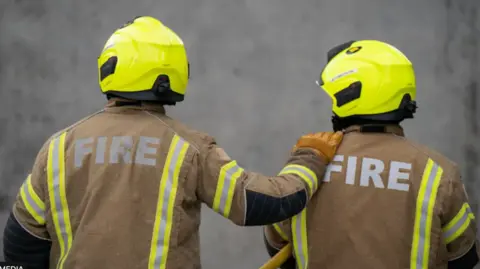 Two firefighters in uniform stand side by side wearing helmets and jackets with yellow hi-vis stripes. One has his hand placed on the other's shoulder. 