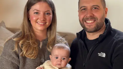 Parents Lizzie and Brad smile into camera holding their baby daughter, Penny. Lizzie has long, dark blonde/brown hair and wears a grey top. Brad has short dark hair and a short dark beard and wears a black jacket.