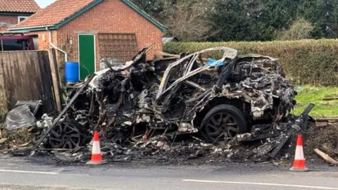 The charred remains of a car. It is beside a road. There are three road cones in front of it. Only the wheels of the car are distinguishable in the blackened wreckage. A garage, a house and the top of another vehicle behind a fence can also be seen.