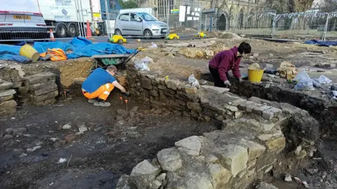 University of Leicester Archaeological Services People digging in the dirt next to the remains of a stone building.