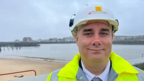 Andrew Turner/BBC Councillor Daniel Candon, wearing a white hard hat and fluorescent yellow jacket, shirt and tie. He is smiling, standing near to the Spending Beach on the River Yare at Great Yarmouth's South Denes, with Gorleston Pier Hotel in the backdrop.