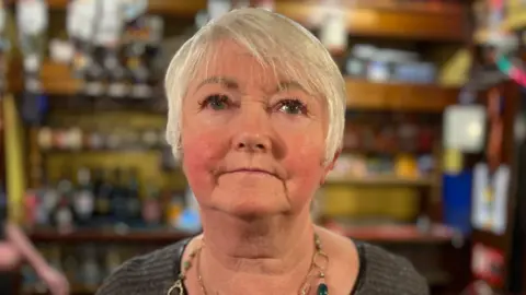 A woman with short blond hair with bar behind.