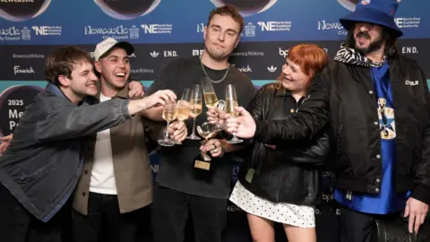 PA Media Sam Fender and his band clinking champagne flutes after the award win. Fender is holding his trophy as well as his own glass.