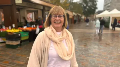 Wendy Randal in a beige jumper, white top and beige scarf, stood in a market