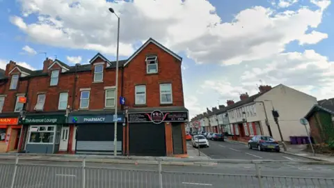 Google streetview showing shops in terraced redbrick properties on the road and other houses on the other side.