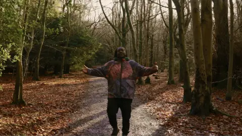 Surrey Hills National Landscape A man, centre, in a forest. He is wearing a burgundy and purple coat and black jeans. He is walking on a muddy path which has green trees either side and orange leaves on the floor. 
