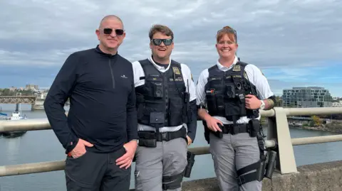 A man wearing sunglasses and a dark-coloured top is standing next to two police officers wearing flak jackets. They are leaning against railings above a body of water with boats and buildings 