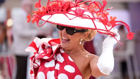 PA Media A woman wears a wide-brimmed red and white hat and red dress with large white dots and smiles