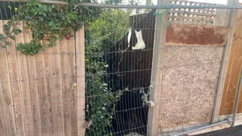 The entrance to a footpath, with overgrown brushes and a gate closing it off. The remnants of a closure sign can be seen tied to the gate. 