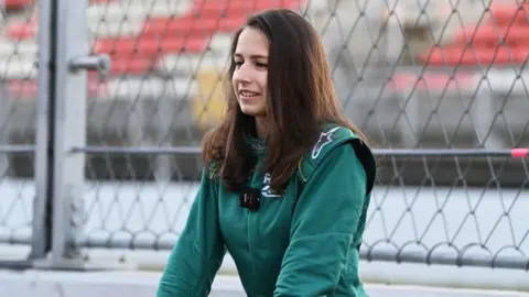 Getty Images Megan Gilkes in a green race suit in front of a chain link fence