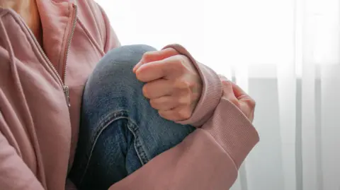 A woman, sitting in front of a window, is clutching her knee to her chest in a supportive gesture. She is wearing a pink zip-up hoodie and blue jeans. She is slightly off centre of the picture and you cannot see her head.