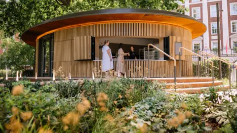 Grosvenor A circular wooden kiosk clad in vertical timber slats, featuring a flat, overhanging roof. Three people stand at the open counter service window. The kiosk is accessed by brick steps with a metal handrail, with green plants in the foreground and a brick building in the background.
