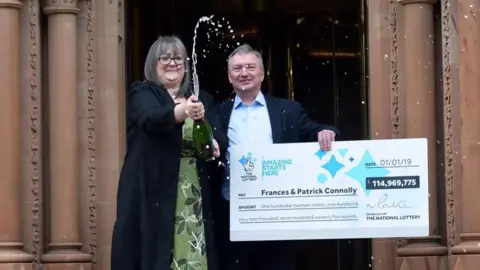 Pacemaker A woman pops open a bottle of champagne as she stands on stpes alongside a mn holding an oversized cheque. He is wearing a blue shirt and jacket. She is wearing a long black coat and green dress. They have won the lottery.