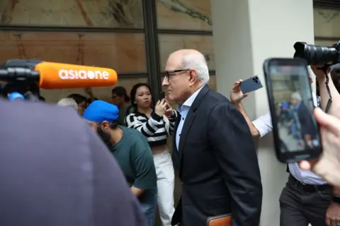 Getty Images Subramaniam Iswaran in glasses and a suit, surrounded by members of the press and people with iPhones