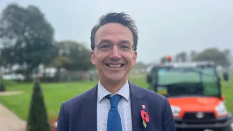 BBC Kirsten Morel is a man wearing glasses, a navy suit jacket with a checked shirt and blue checked tie. He is wearing a jersey pin and a paper poppy on this left lapel. He is smiling into the camera. The background is an out of focus field with trees and an orange tractor.