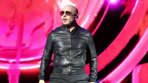 Getty Images Pitbull performs on stage, wearing a black bomber jacket and black sunglasses.