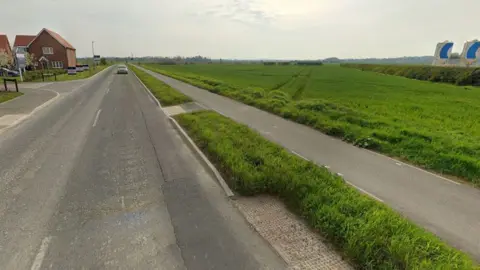 A view of the proposed site on the right side of a road and cycle path/pavement. There is a large field bordered by a hedge on one side. The road is on the left of the picture next to a housing estate on the edge of the village. 