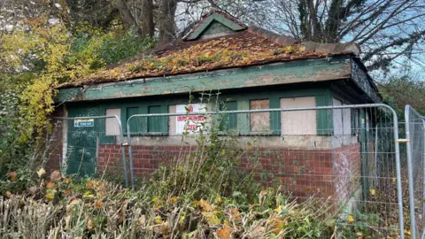 Durham County Council The old toilet block is a one story brown-brick building with a triangle roof and green door and window panes. There is a pile of yellow tree leaves on the roof. NO UNAUTHORISED ACCESS is written in red on a sign attached to the building. A grey metal fence is surrounding the block.
