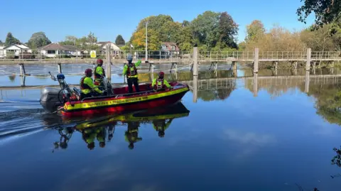 A boat with four rescue workers in it. They are on a weir, with trees in the background.